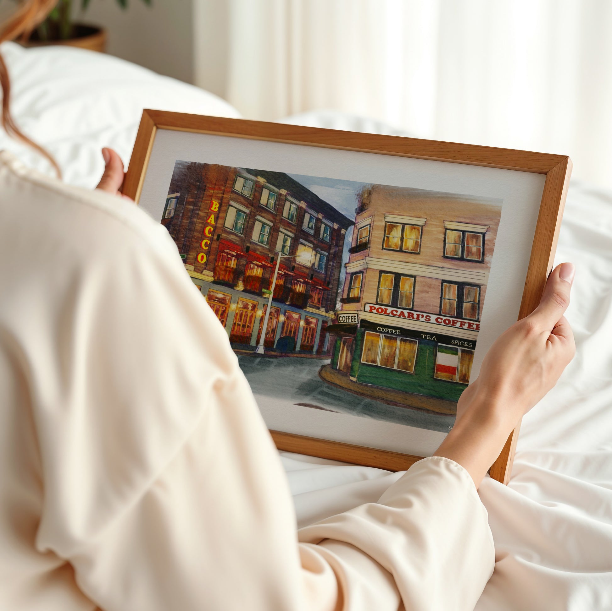 A person in a cream-colored robe holds a framed watercolor painting of a street scene with colorful buildings. The photo is taken from behind the person, showing their hands holding the square wooden frame against a soft, white bedroom background.