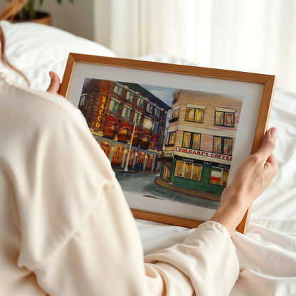 A person in a cream-colored robe holds a framed watercolor painting of a street scene with colorful buildings. The photo is taken from behind the person, showing their hands holding the square wooden frame against a soft, white bedroom background.