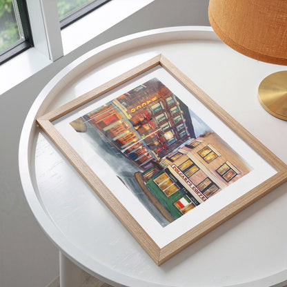 A framed art print depicting a colorful street scene with buildings and storefronts, displayed on a white round table next to a lamp.