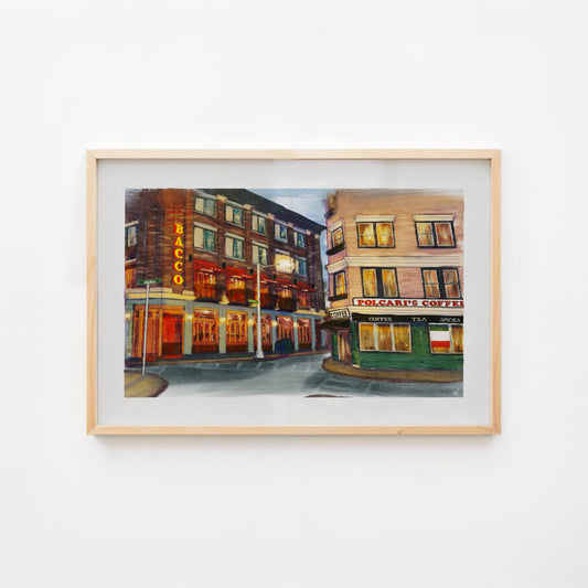 A framed watercolor painting of a city street corner with brick buildings, storefronts, and warm glowing windows, displayed against a plain white wall.