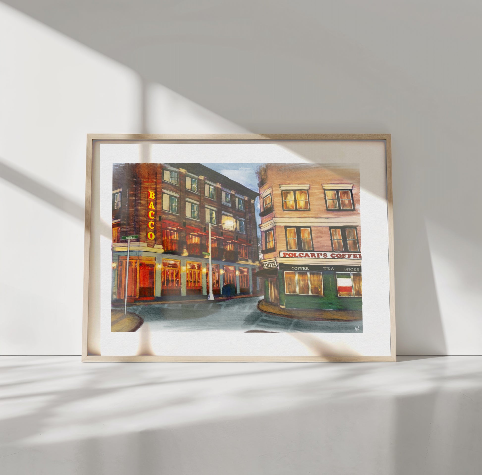 A framed art print depicting a vintage street scene with brick buildings, warm glowing windows, and storefronts, displayed against a white wall with soft shadow patterns.
