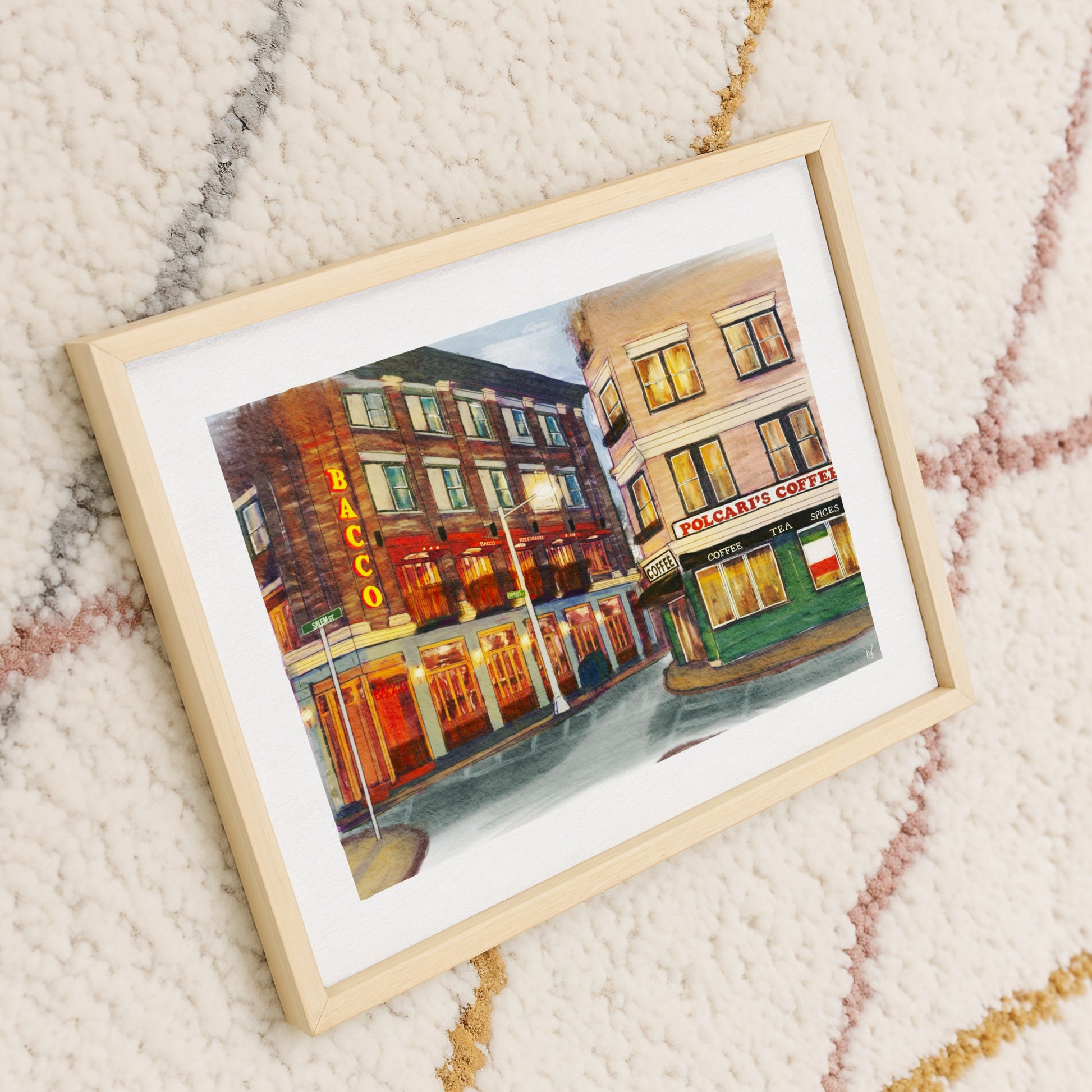 A framed watercolor painting of a city street scene with storefronts, shown at an angle on a textured cream rug with subtle pink and gold patterns. The print features a light wood frame and white mat.