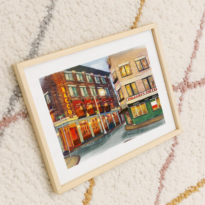 A framed watercolor painting of a city street scene with storefronts, shown at an angle on a textured cream rug with subtle pink and gold patterns. The print features a light wood frame and white mat.