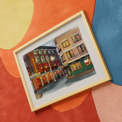 A framed watercolor painting of a street scene with storefronts, shown at an angle on a colorful abstract rug. The artwork depicts buildings with warm glowing windows and signage, including a visible 'Coca-Cola' sign.