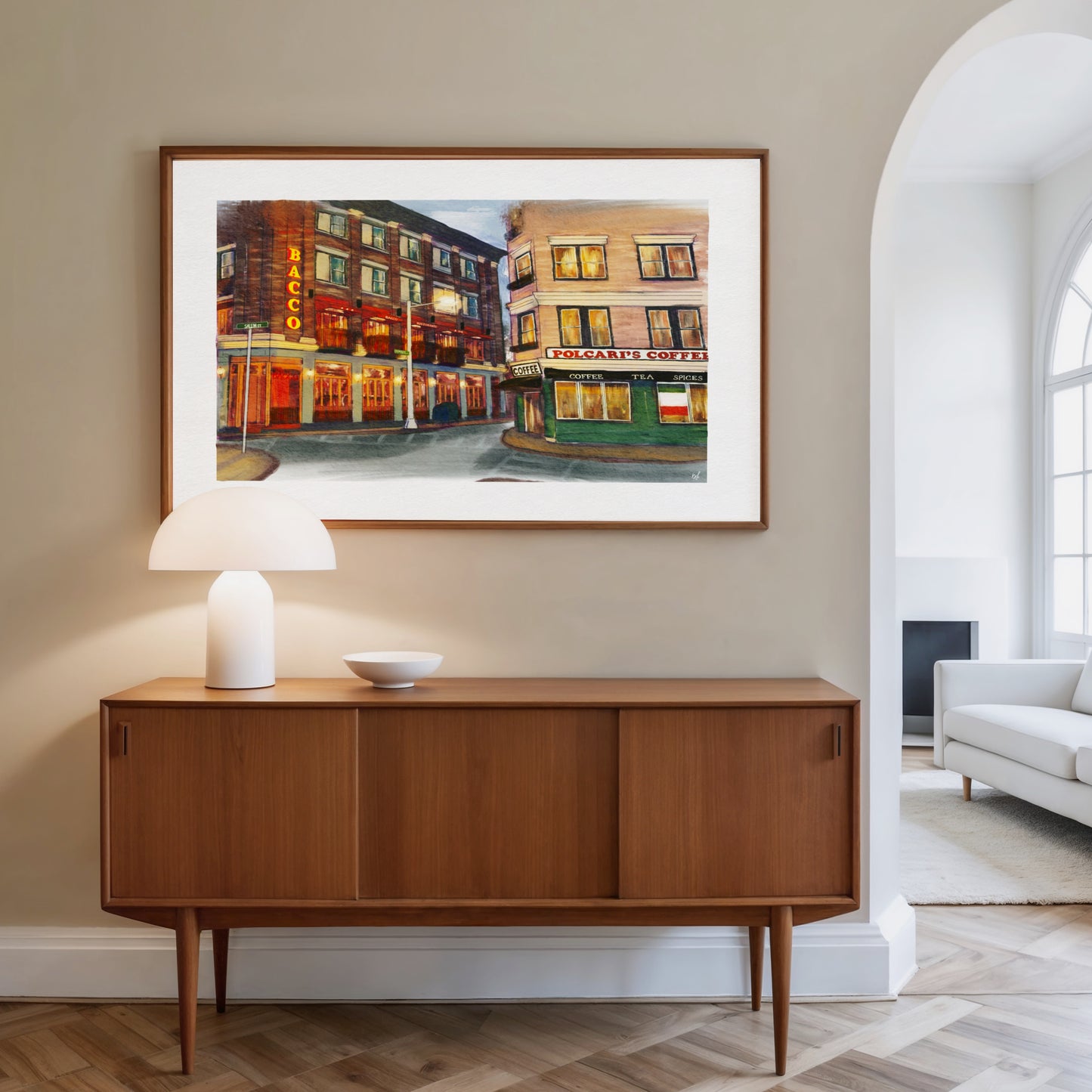 A framed art print depicting a vintage street scene with warm-toned buildings and glowing windows, displayed on a beige wall above a mid-century modern wooden sideboard with a white table lamp.