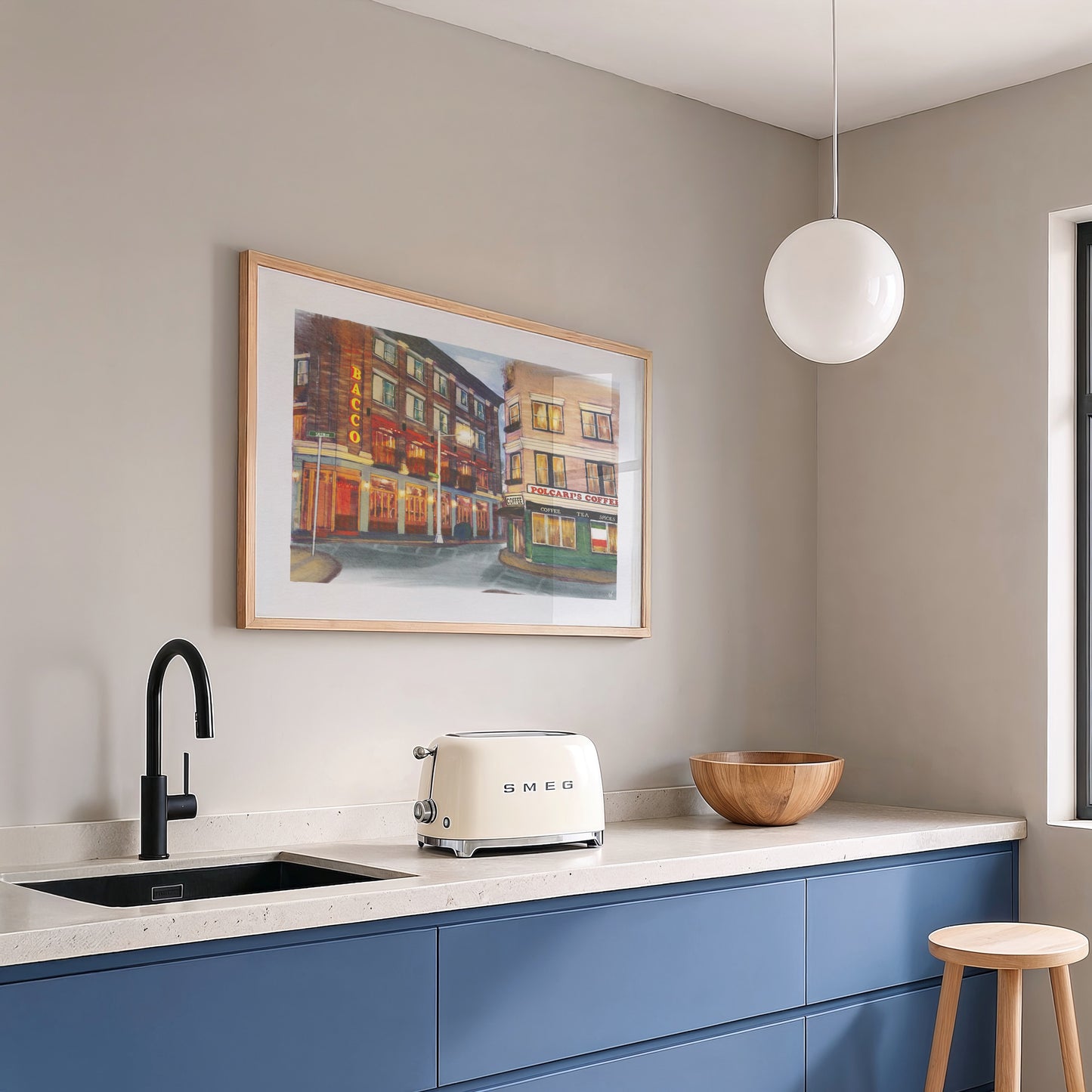 A framed art print depicting a colorful city street scene hangs on a gray wall above a kitchen counter with blue cabinets, a black faucet, a cream Smeag toaster, and a wooden bowl.